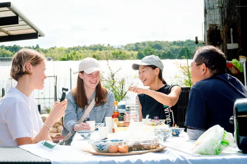 屋外の食卓で、茶碗や卵を前に、4人が食べながら笑って話している